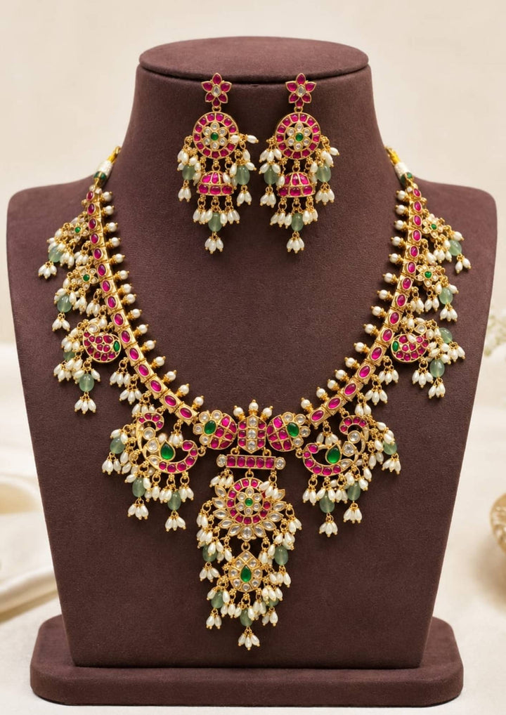 Elegant gold plated Kundan necklace set adorned with pink ruby stones, green accents, floral motifs, pearl drops, and matching earrings for festive occasions.