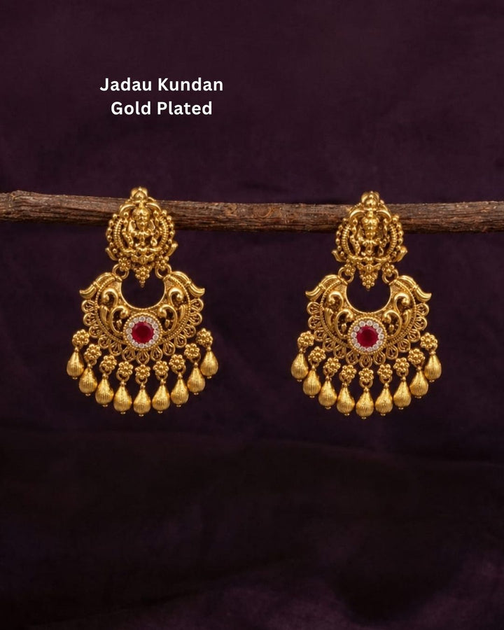 Gold-plated Laxmi Set jewelry necklace and matching earrings displayed on a dark red velvet bust. The necklace features intricate gold filigree, red and green stone accents, and rows of white faux pearls.