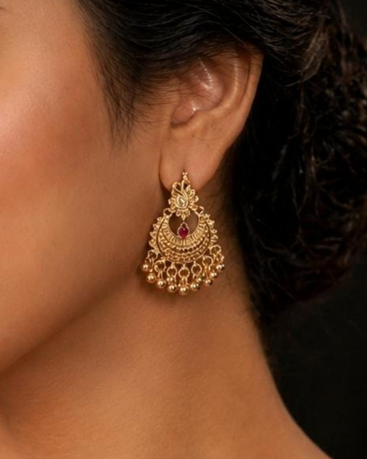 A pair of gold-plated traditional Indian jhumka earrings with intricate filigree detailing and a small red stone accent, hanging from a wooden branch against a dark background. White text in the upper left reads &