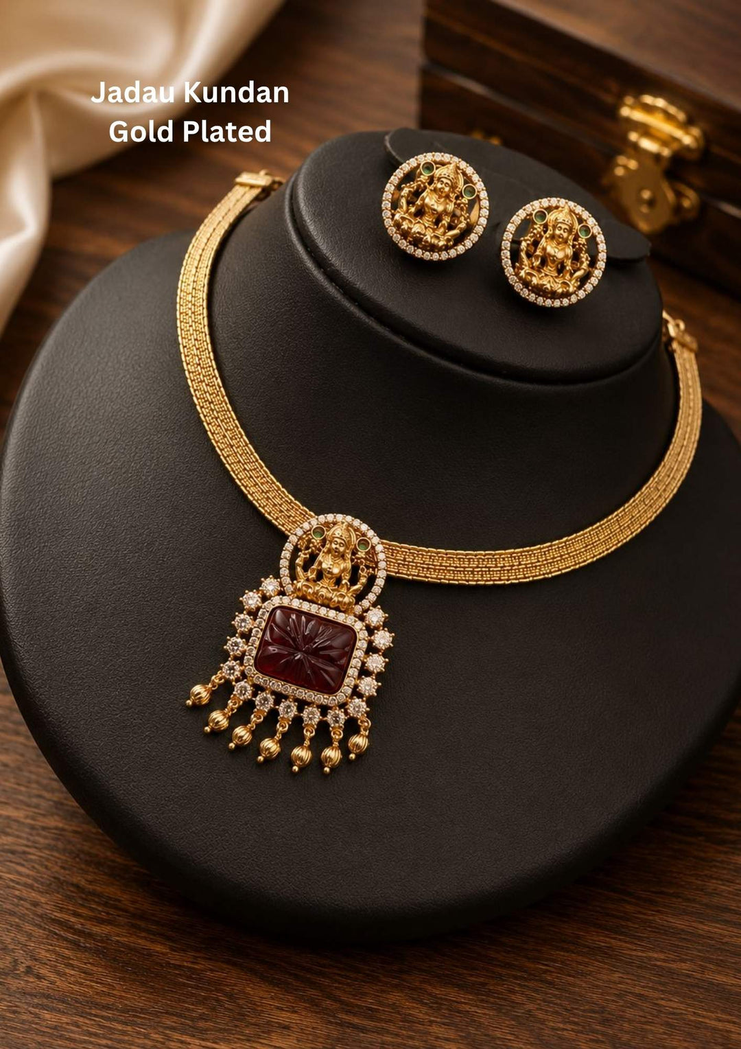 A traditional Jadau Kundan gold plated jewelry set featuring a rigid, textured mesh-style hasli wire collar neckpiece. The center is adorned with a square-cut, intricately carved deep ruby-red stone pendant topped with a detailed miniature figurine of sitting Goddess Lakshmi. The set includes matching round coin-style stud earrings bordered with tiny white crystals.