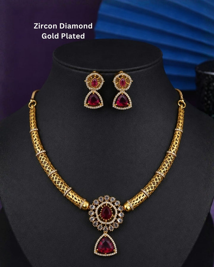 Gold-plated necklace and matching earrings set with red gemstone accents, displayed on a black jewelry bust. The necklace features a textured gold chain and a central pendant with a red teardrop gemstone surrounded by clear stones.