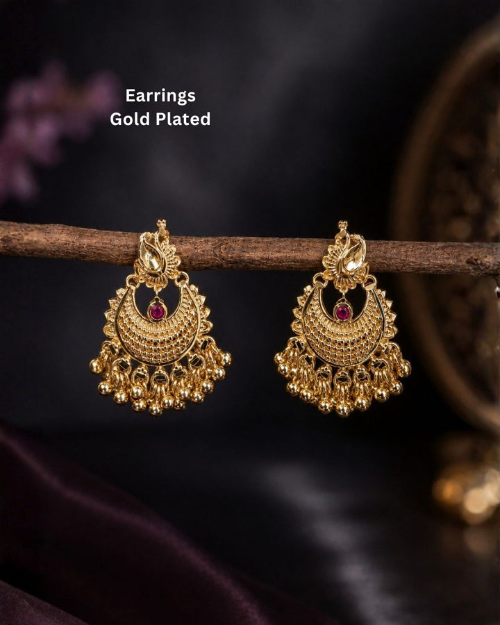 A pair of gold-plated traditional Indian jhumka earrings with intricate filigree detailing and a small red stone accent, hanging from a wooden branch against a dark background. White text in the upper left reads &