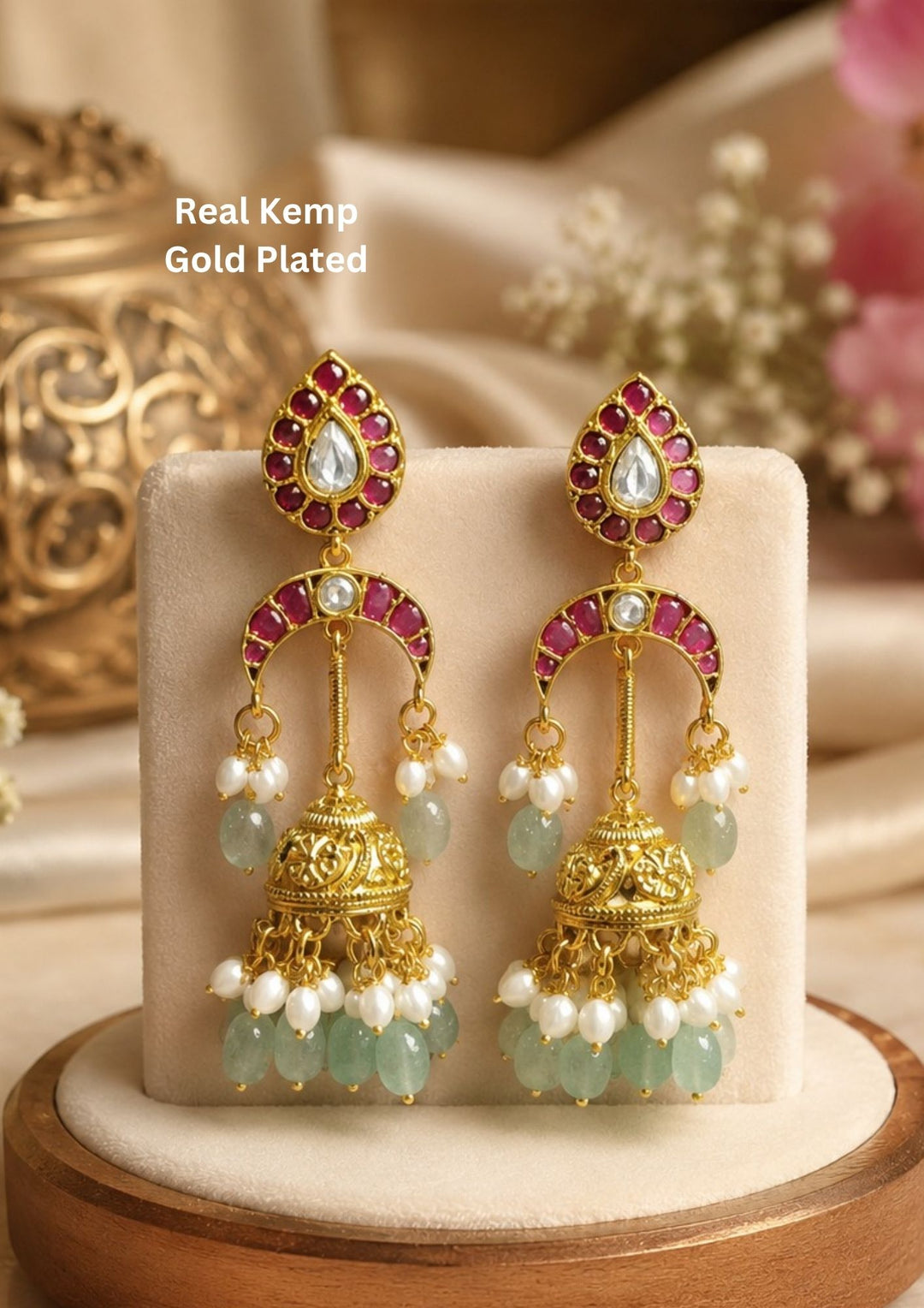 Beautiful ruby stone gold plated jhumka earrings featuring teardrop-shaped stud tops embellished with white kundan stones and ruby accents. The elegant crescent design connects to intricately crafted dome-shaped jhumkas adorned with pearl clusters and green gemstone bead danglers for a rich traditional look.
