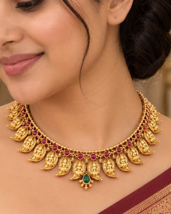 Elegant gold plated Lakshmi Devi necklace set featuring multiple detailed temple motifs, red and green stone embellishments, a central green teardrop pendant, and matching earrings, perfect for festive and traditional occasions.