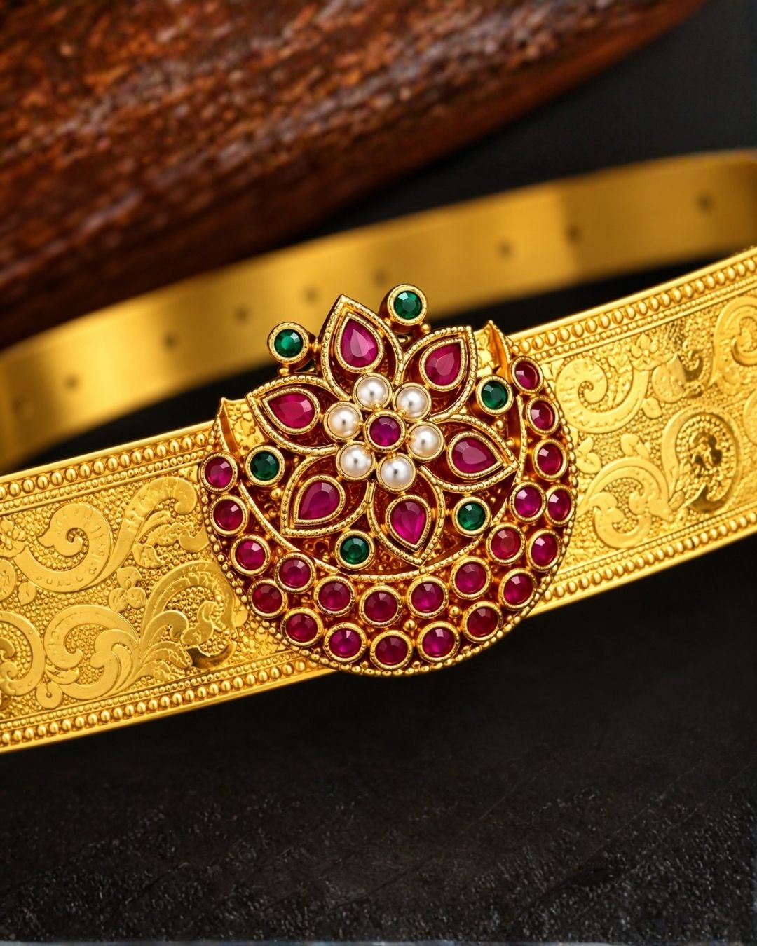 Beautiful gold plated hip belt featuring an intricately engraved wide band with a floral gemstone centerpiece accented with ruby, green, and white stones. Perfect for traditional wear, weddings, festive occasions, bridal styling, parties, and gifting.