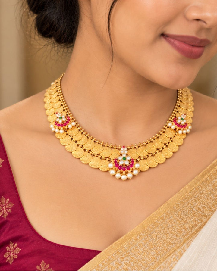 Traditional gold plated designer necklace and earrings set featuring rows of detailed coin motifs, floral designs with ruby, green and white stones, and pearl drops, perfect for festive, wedding, and traditional occasions.