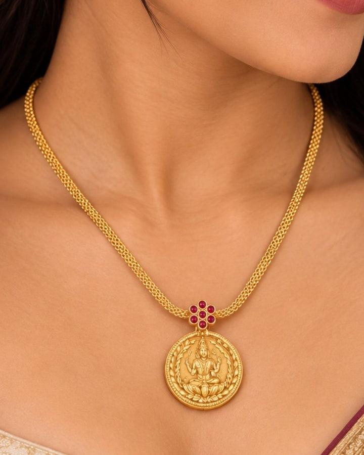 Woman wearing a gold necklace with a coin pendant Laxmi chain gold plated traditional necklace featuring a circular coin pendant embossed with Goddess Lakshmi seated on a lotus, connected by a small pink ruby stone floral accent to a thick textured mesh chain. This classic South Indian style necklace is crafted with a premium gold finish that provides a rich divine appearance suitable for weddings, festivals, poojas, family functions, Varalakshmi Vratam, Diwali, sarees, and ethnic outfits with skin-friendly