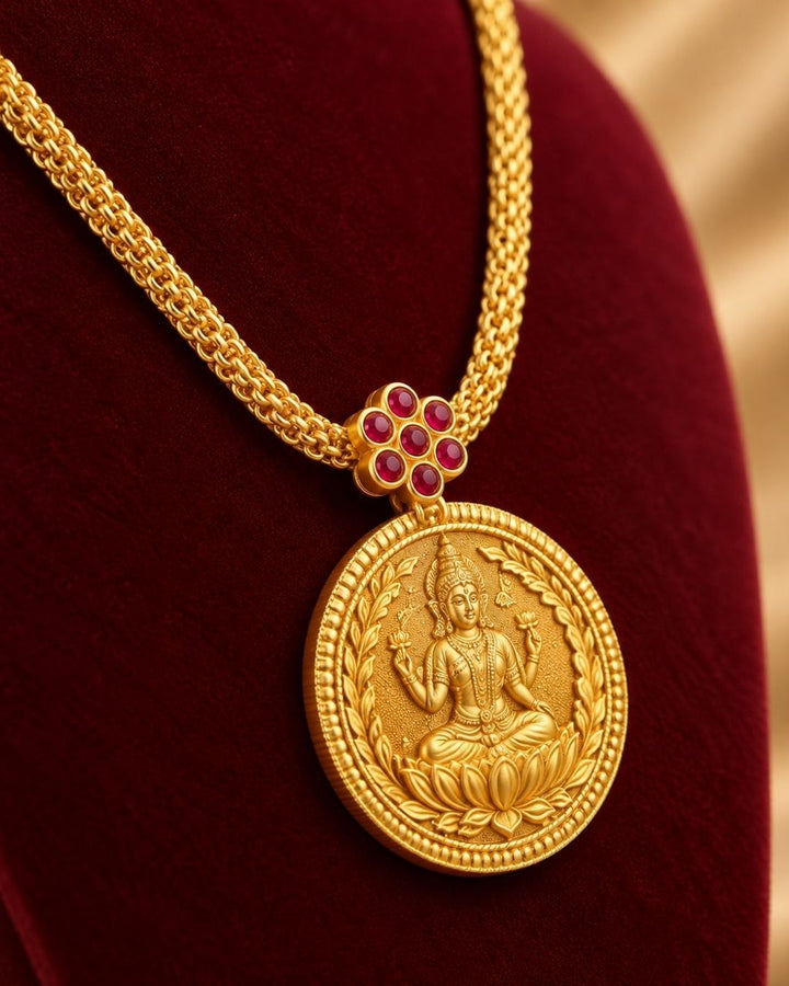 Woman wearing a gold necklace with a coin pendant Laxmi chain gold plated traditional necklace featuring a circular coin pendant embossed with Goddess Lakshmi seated on a lotus, connected by a small pink ruby stone floral accent to a thick textured mesh chain. This classic South Indian style necklace is crafted with a premium gold finish that provides a rich divine appearance suitable for weddings, festivals, poojas, family functions, Varalakshmi Vratam, Diwali, sarees, and ethnic outfits with skin-friendly