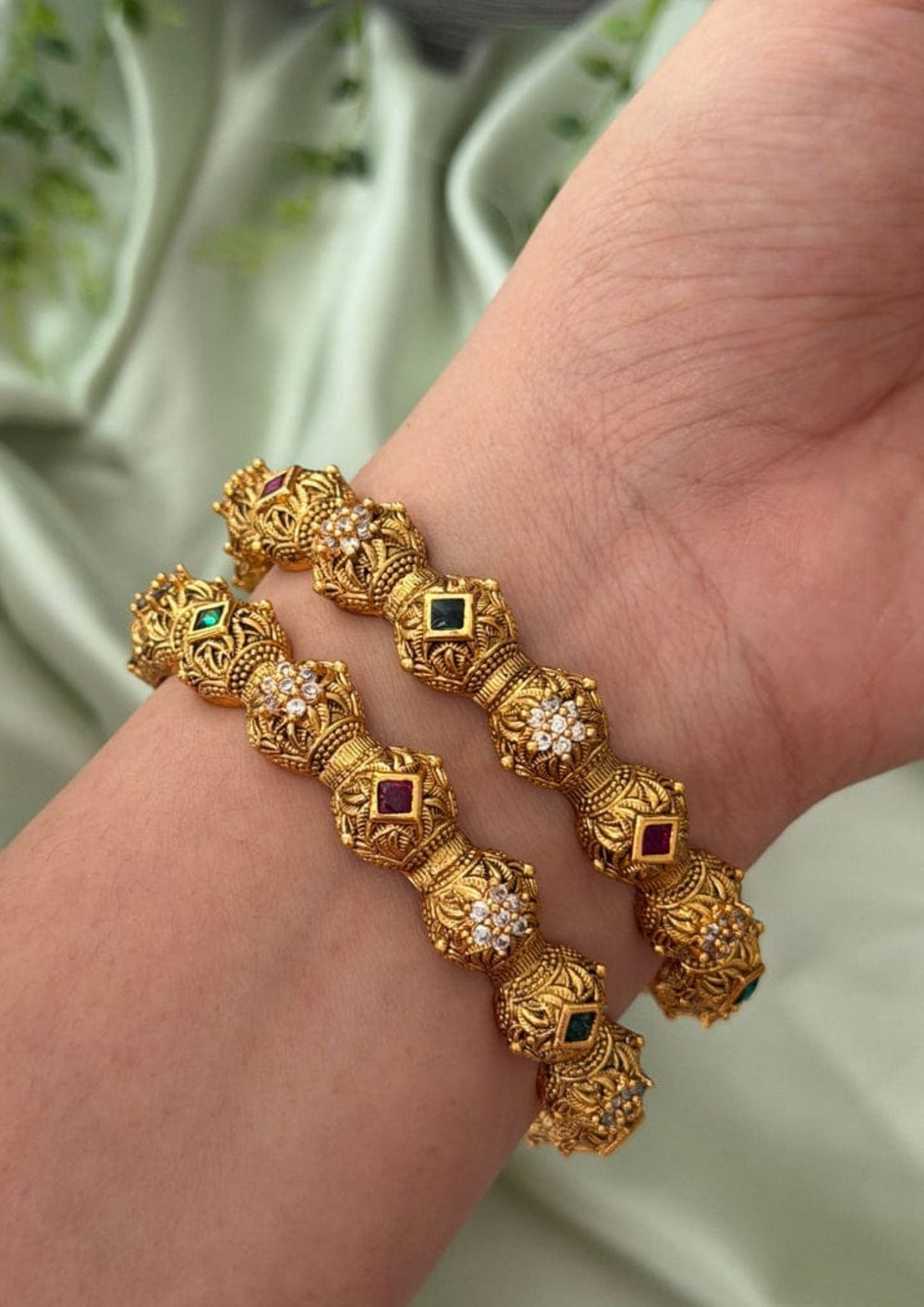 Close up of a south indian women hand wearing Traditional gold plated temple bangles showcasing ornate antique detailing, ruby pink square stones, emerald green accents, and floral zircon motifs. Perfect for bridal wear, festive occasions, and ethnic celebrations, offering a luxurious heritage-inspired look.