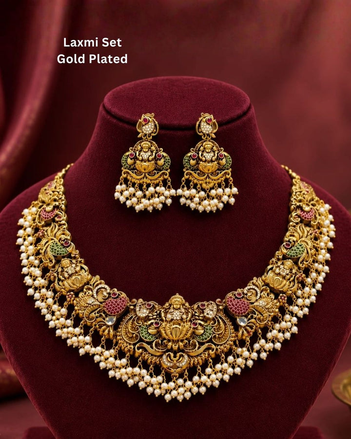Elegant antique gold plated temple choker necklace adorned with Lakshmi and peacock motifs, ruby and green stone accents, pearl drops, and matching earrings.