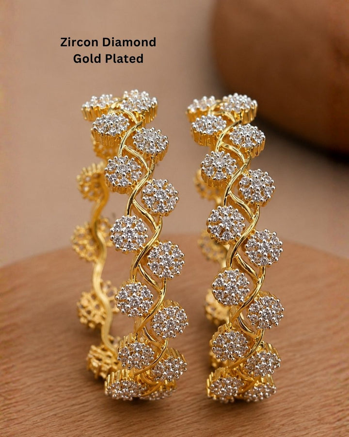 A pair of gold-plated huggie bangles decorated with clear zircon stones in a floral pattern, displayed on a wooden surface with a warm brown background. Text overlay reads &