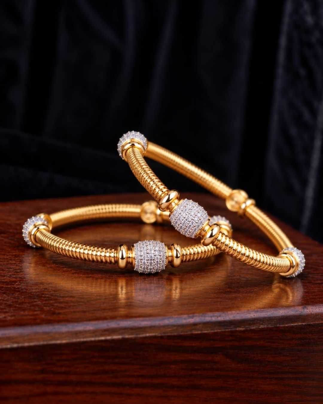 Pair of gold plated designer bangles displayed on a dark wooden surface against a black fabric background.