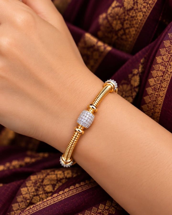 Pair of gold plated designer bangles displayed on a dark wooden surface against a black fabric background.
