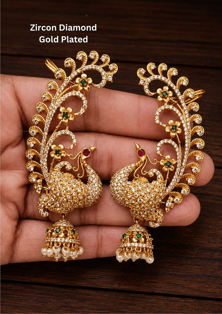 Gold plated Zircon Diamond ear cuff earrings designed like a peacock tail plume decorated with white CZ stones, small gold branches, and green-centered flower details. The bottom has an embossed peacock design with white stones and a red stone eye, from which a hanging dome jhumka is attached featuring green stone details and white pearl drops along the bottom rim, providing a premium heritage appearance suitable for family gatherings, festivals, parties, and weddings with anti fade color