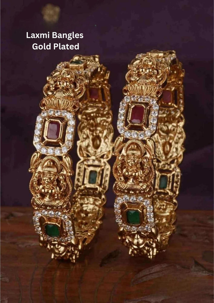 A pair of heavy, antique 18k gold plated temple kadas featuring repeating embossed panels of Goddess Lakshmi seated on a lotus throne. Each sacred idol frame is bordered with fine white zircon stones and interspersed with rectangular settings containing alternating ruby red and emerald green gemstones.