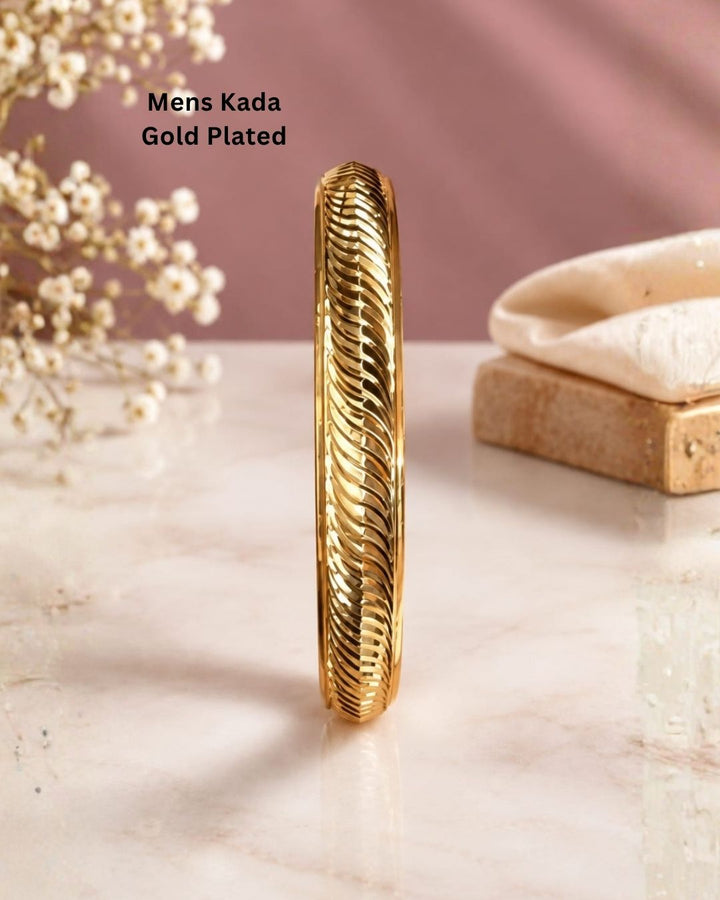 Gold plated men’s kada displayed upright on a beige surface with a soft pink background, highlighting its woven wave design and polished finish.