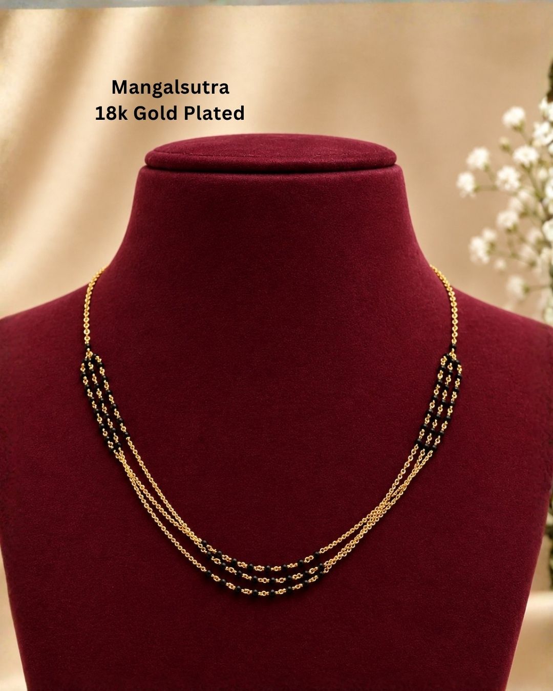 A Mangalsutra necklace made of 18k gold-plated metal with black and gold beadwork, displayed on a dark red velvet jewelry bust. The text &