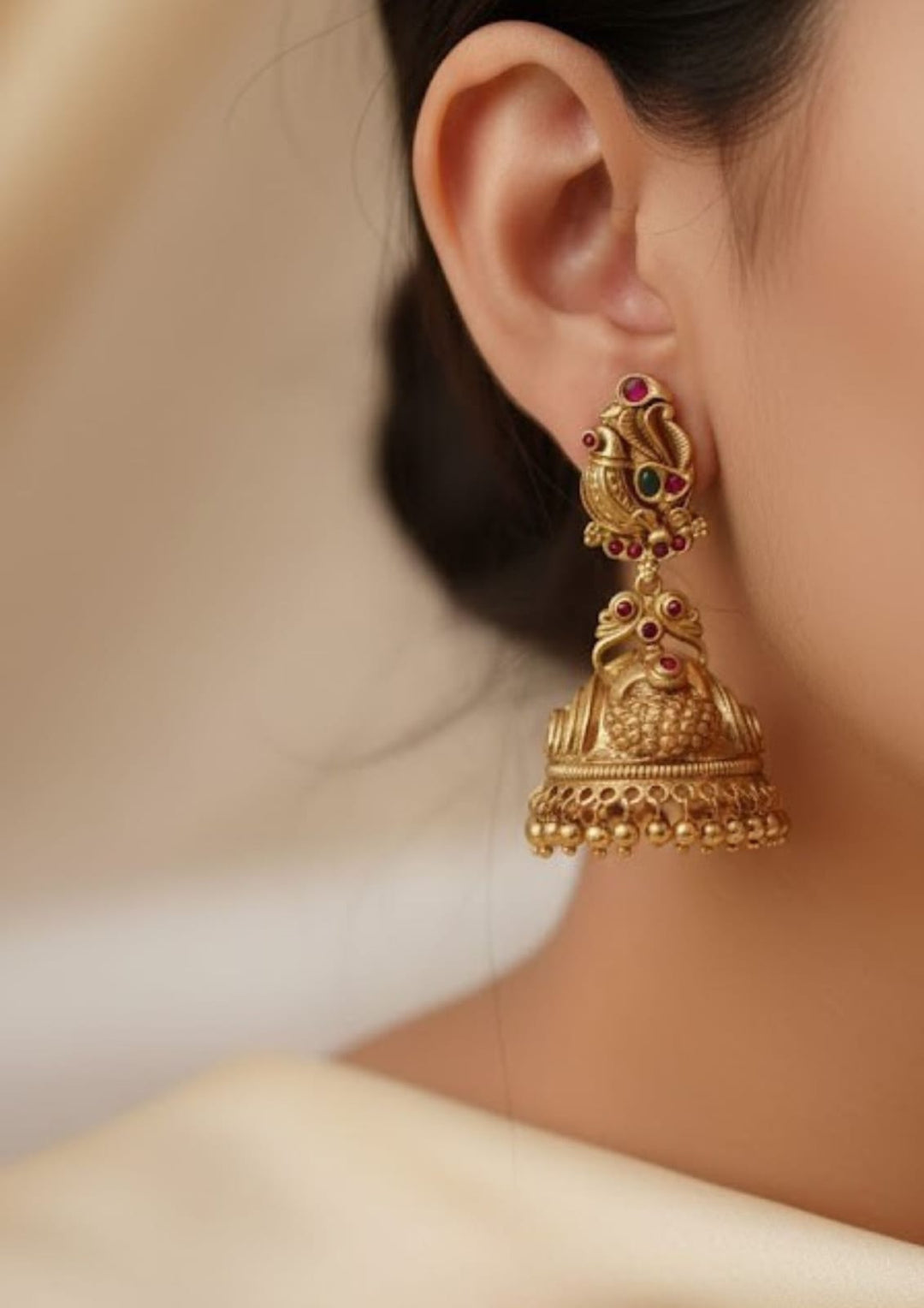 close up of south indan women wearing a Gold plated Jadau Kundan jhumka earrings designed with peacock top stud decorated with small ruby pink and emerald green stone accents. The traditional antique dome-shaped jhumki base is crafted with Nakshi floral engravings, dainty colored stone highlights, and finished with a dense, multi-layered of hanging gold beads suitable for family gatherings, festivals, parties, and weddings with skin friendly
