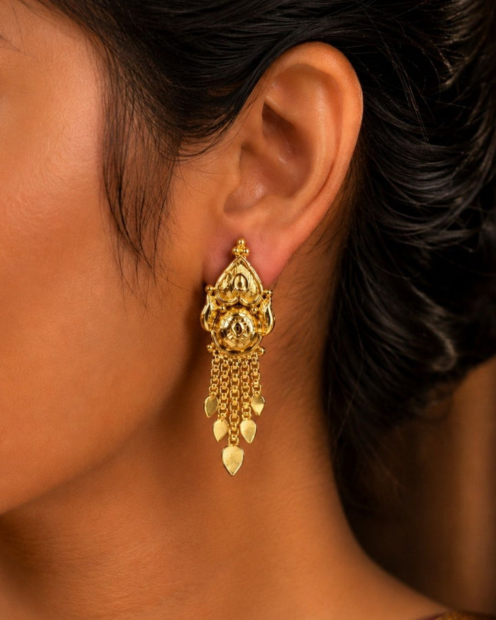 A pair of gold-plated traditional Indian jhumka earrings hanging from a rustic wooden branch against a solid black background, with white text overlay reading &