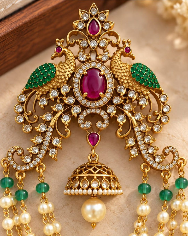 Gold plated traditional hair accessory featuring an intricate peacock-inspired design, ruby red stones, green accents, sparkling white stones, and cascading pearl chains.