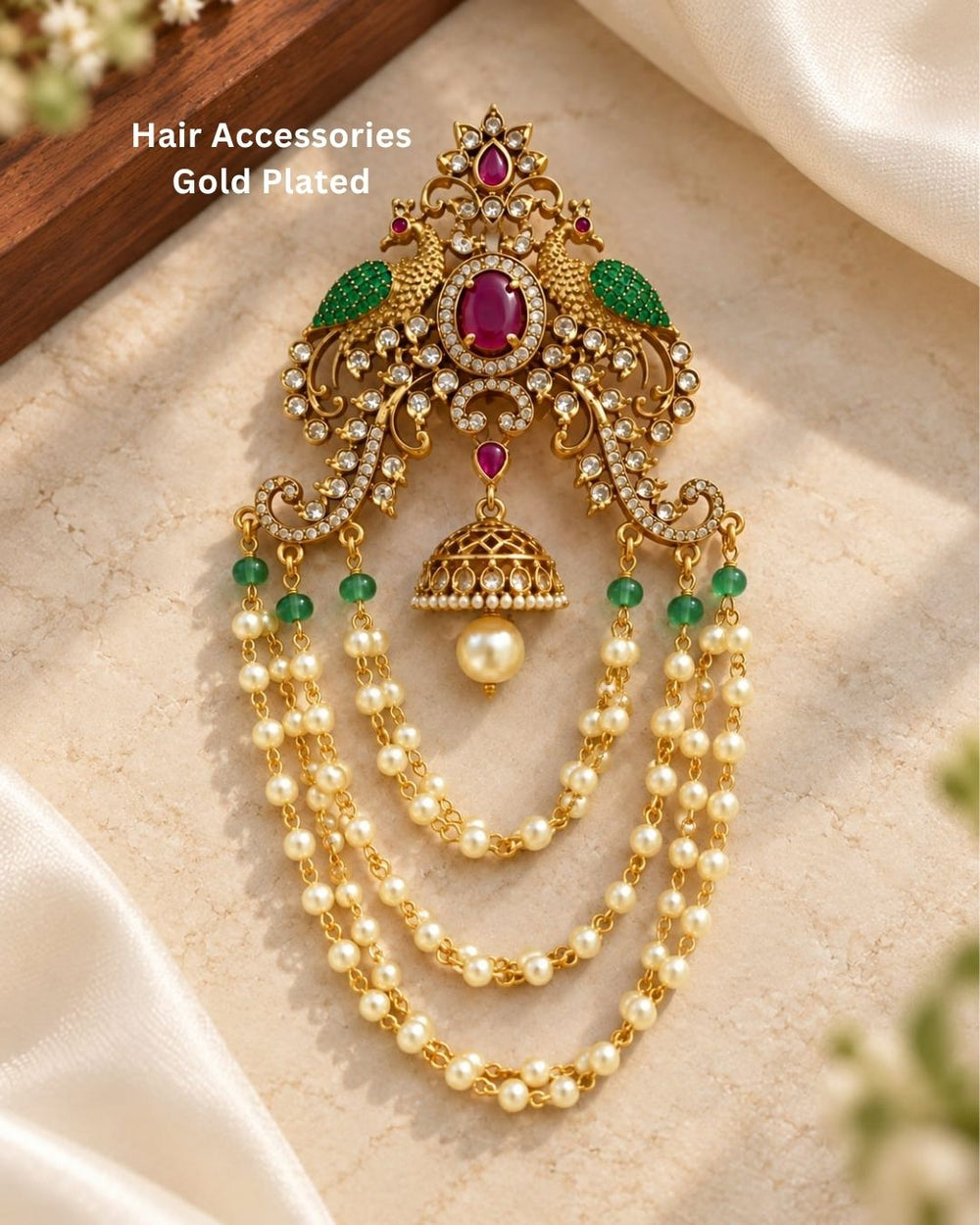 Gold plated traditional hair accessory featuring an intricate peacock-inspired design, ruby red stones, green accents, sparkling white stones, and cascading pearl chains.