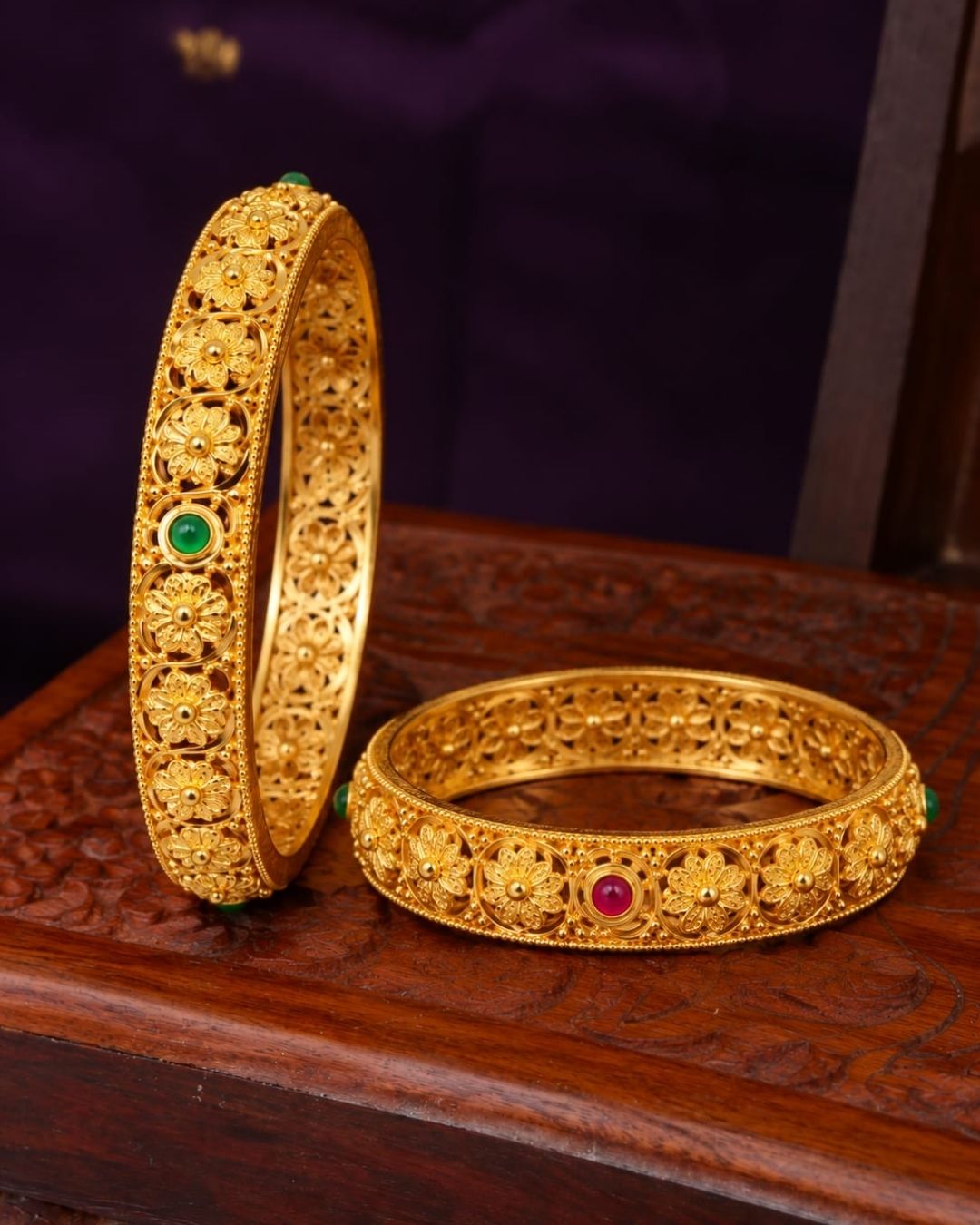 Elegant 18K gold plated floral bangles displayed on a carved wooden jewellery box, showcasing ornate gold craftsmanship and green and ruby stone details.