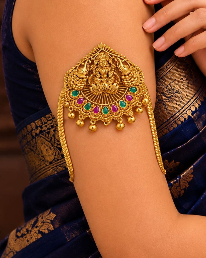 Traditional gold plated designer vanki featuring a detailed Goddess Lakshmi centerpiece, peacock motifs, alternating green and ruby-colored stones, intricate temple-inspired detailing, and hanging gold beads, perfect for festive and wedding occasions.