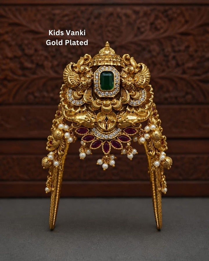 Beautiful gold plated kids vanki featuring an intricately crafted traditional design with a large green stone centerpiece, sparkling clear stones, ruby floral motifs, and elegant pearl drops. Perfect for traditional wear, weddings, festivals, classical dance, special occasions, and gifting.