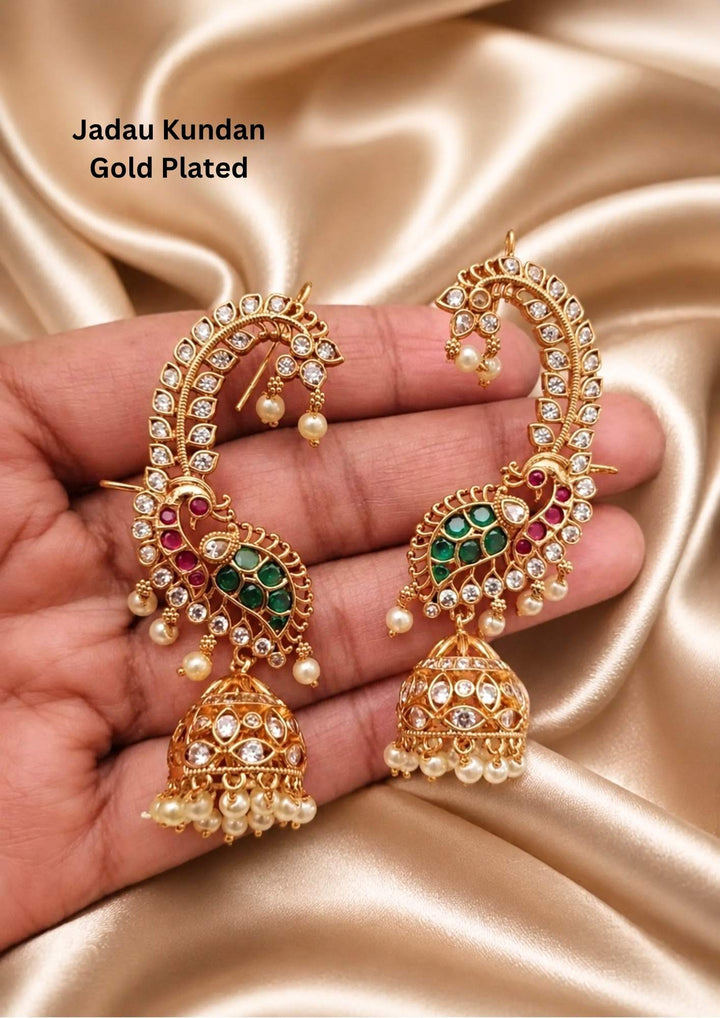 Traditional Jadau Kundan 18k gold plated ear cuff earrings or chain jhumkas. The unique design features a long, curved gold cuff shaped like a peacock's neck that hugs the ear contour, encrusted with white kundan stones and hanging small pearls. The base features a detailed peacock motif filled with ruby pink and emerald green stone inlay, from which a classic dome-shaped jhumki hangs with a cluster of white pearl drops. Perfect for weddingwear and it is anti fade color.