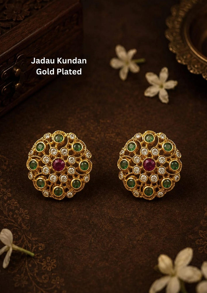 Traditional jadau kundan gold plated stud earrings featuring an intricate circular floral design crafted with radiant emerald-colored stones, sparkling kundan stones and a striking pink center stone. The symmetrical pattern and antique-inspired craftsmanship create a rich ethnic appeal suitable for weddings, festivals, poojas and traditional celebrations.