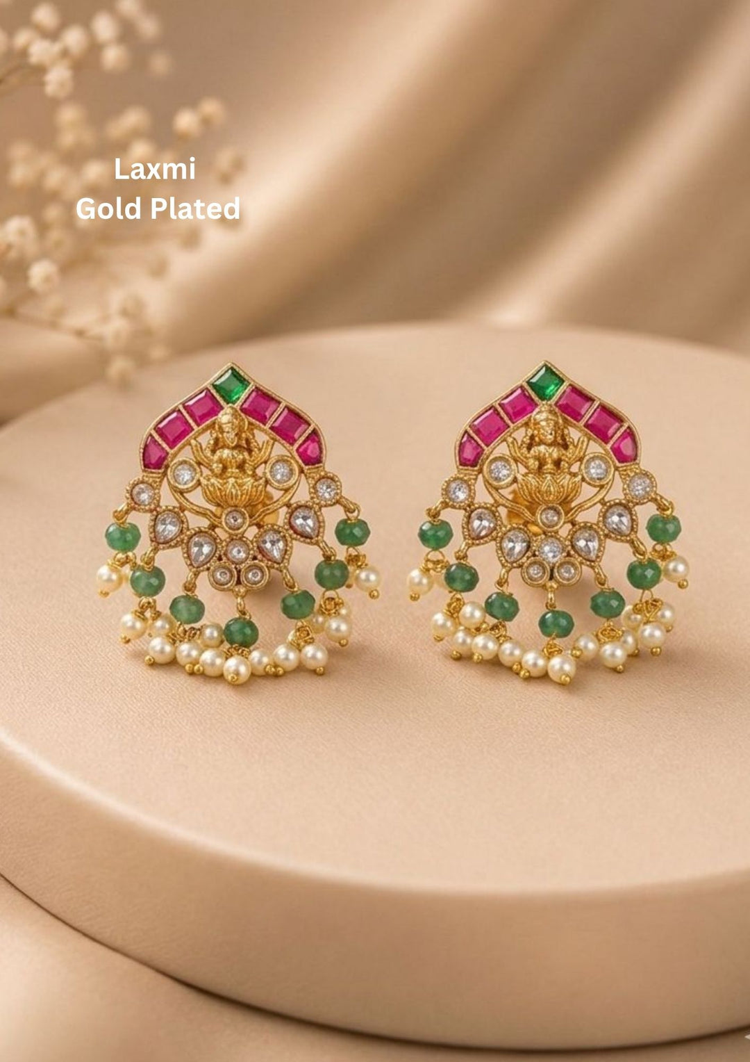 A pair of antique temple-style gold-plated stud earrings intricately crafted with a central Goddess Laxmi motif seated on a lotus flower. The top edge is bordered with an arch of square-cut pink kemp stones and a single green accent, while the lower filigree tier is adorned with small white stones, hanging emerald-green round beads. Perfect for weddings and festive wear.