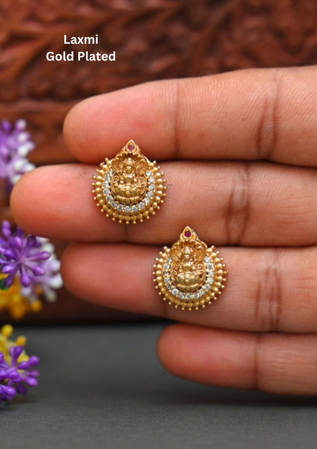Traditional Laxmi gold plated stud earrings . The circular ear studs feature a highly detailed embossed figure of Goddess Lakshmi sitting on a lotus in the center. The idol is semi-circled by shimmering white CZ American diamond stones, a beaded gold border, and a small red ruby stone at the very top tip. This temple jewelry piece provides an elegant ethnic look suitable for puja festivals, weddings, and traditional family occasions. 
