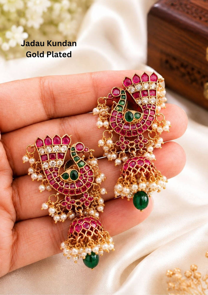 jadau kundan gold plated jhumka earrings featuring an intricate peacock motif studded with pink ruby stones, green stone accents, and clear kundan detailing. These lightweight traditional earrings are crafted with a classic dome-shaped jhumka base adorned with dangling white pearl clusters and a smooth green drop bead at the bottom, offering a rich royal appearance suitable for weddings, festivals, poojas, sarees, lehengas, and ethnic outfits with skin-friendly quality, anti-fade polish, comfortable fitting