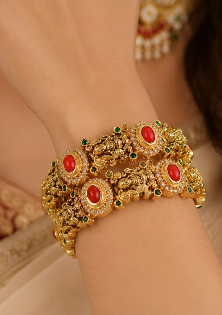 Traditional South Indian woman wearing temple design coral stone gold plated bangles. The bangles feature detailed Lakshmi-inspired carvings, red oval stones, green accents, and antique gold finish, making them ideal for weddings, festivals, and bridal wear.