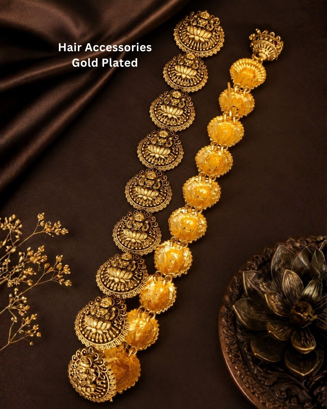 A pair of gold-plated traditional Indian hair accessories, featuring a long chain of ornate circular jhumka-style beads and a matching floral garland, displayed on a dark brown satin fabric background with decorative dried flowers.