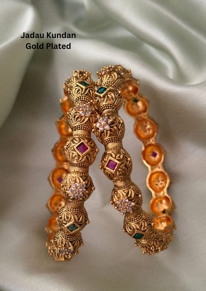 Elegant temple design gold plated bangles featuring square ruby pink and emerald green stones set amidst intricate antique floral carvings. Adorned with sparkling white zircon clusters and detailed handcrafted patterns, these bangles reflect timeless South Indian temple jewelry artistry.