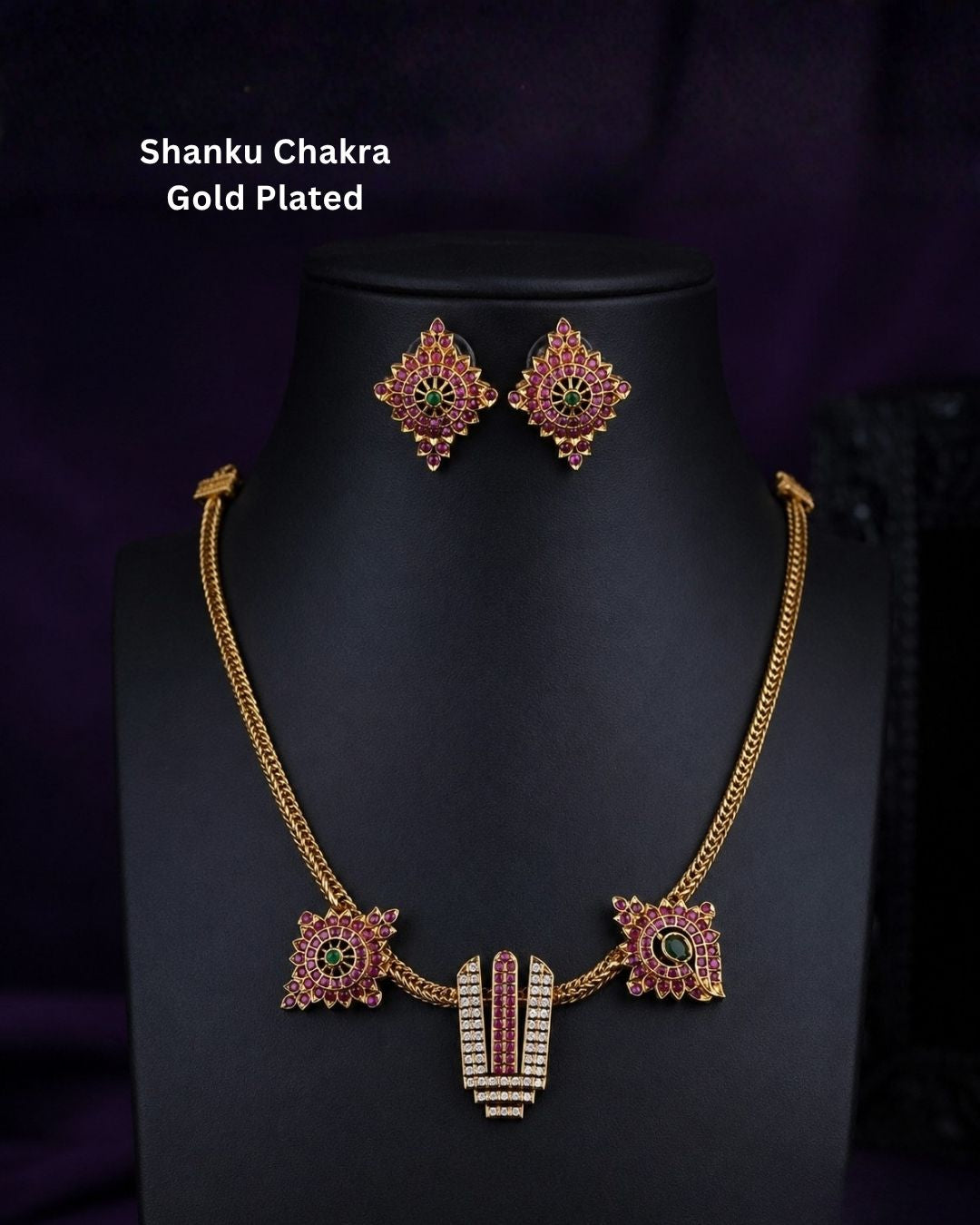 Gold-plated Shanku Chakra necklace and matching earrings displayed on a black mannequin bust against a dark purple background. The necklace features a central vertical Shanku Chakra pendant with red and green stone detailing, flanked by two matching square earrings.