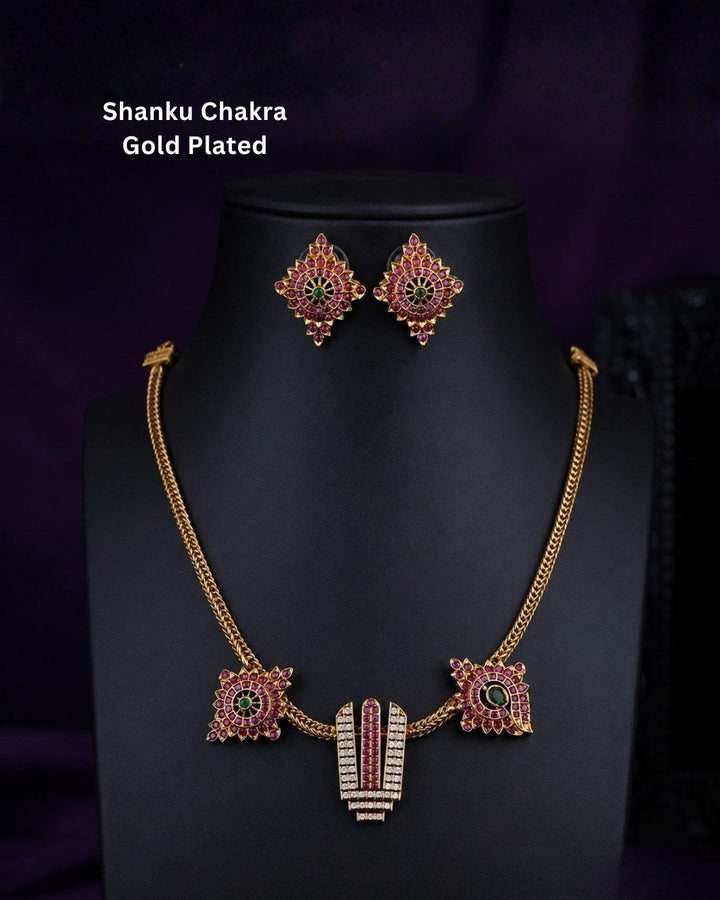 Gold-plated Shanku Chakra necklace and matching earrings displayed on a black mannequin bust against a dark purple background. The necklace features a central vertical Shanku Chakra pendant with red and green stone detailing, flanked by two matching square earrings.