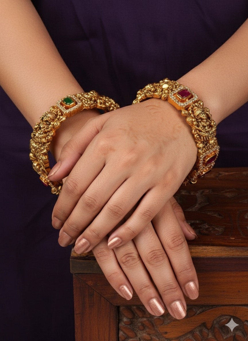 south indian women wearing gold plated temple kadas featuring repeating embossed panels of Goddess Lakshmi seated on a lotus throne. Each sacred idol frame is bordered with fine white zircon stones and interspersed with rectangular settings containing alternating ruby red and emerald green gemstones.