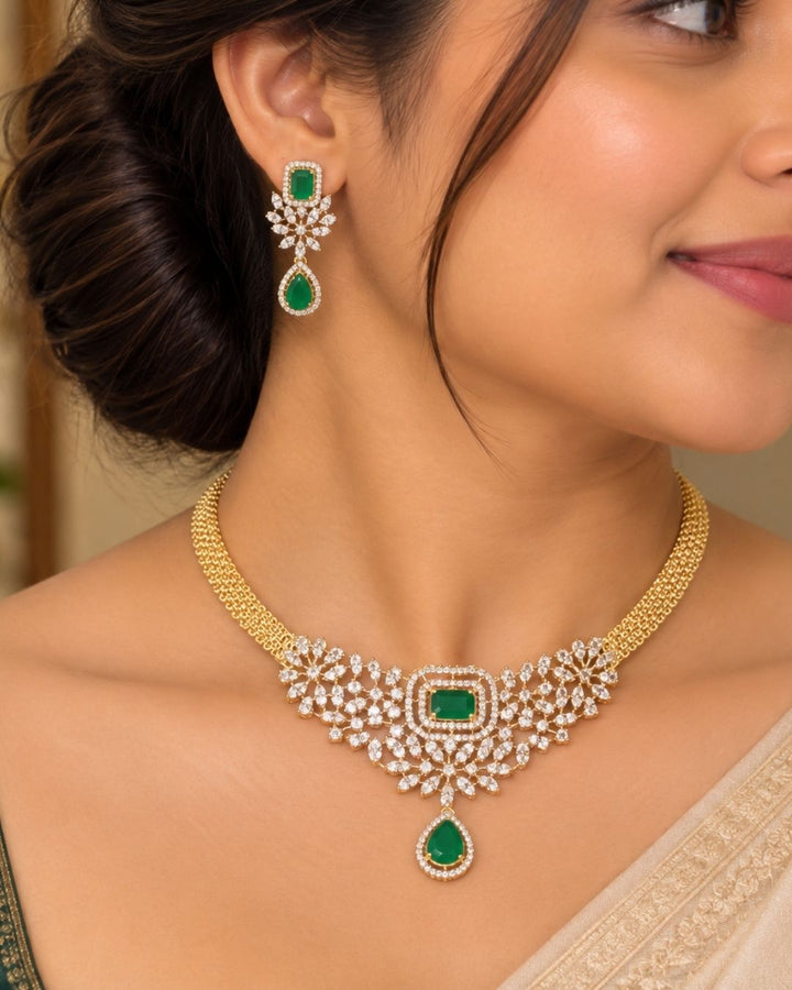 It centers an intricate, V-shaped floral bib pattern encrusted with glittering white American Diamonds (CZ stones), a prominent square emerald-green center stone, and a dangling teardrop emerald charm at the base. Includes a matching pair of elegant hanging earrings featuring a floral design with square and teardrop green stone accents. Perfect for parties and festive occasions with skin-friendly premium quality, comfortable fit,