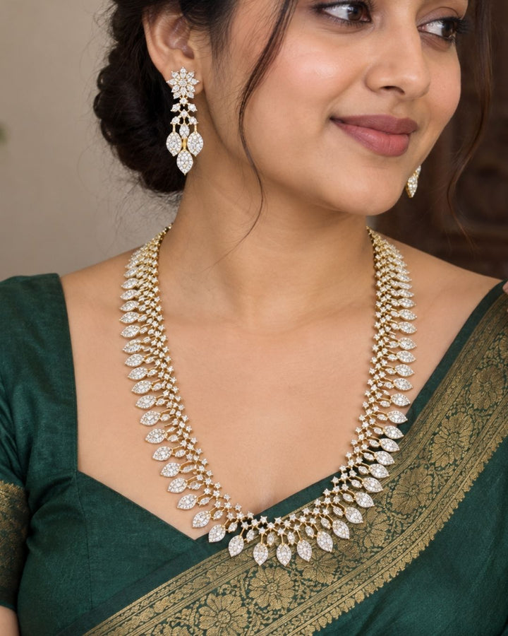 Designer gold plated long necklace and matching earrings featuring intricate leaf motifs embellished with sparkling white stones, perfect for festive, wedding, and traditional occasions.