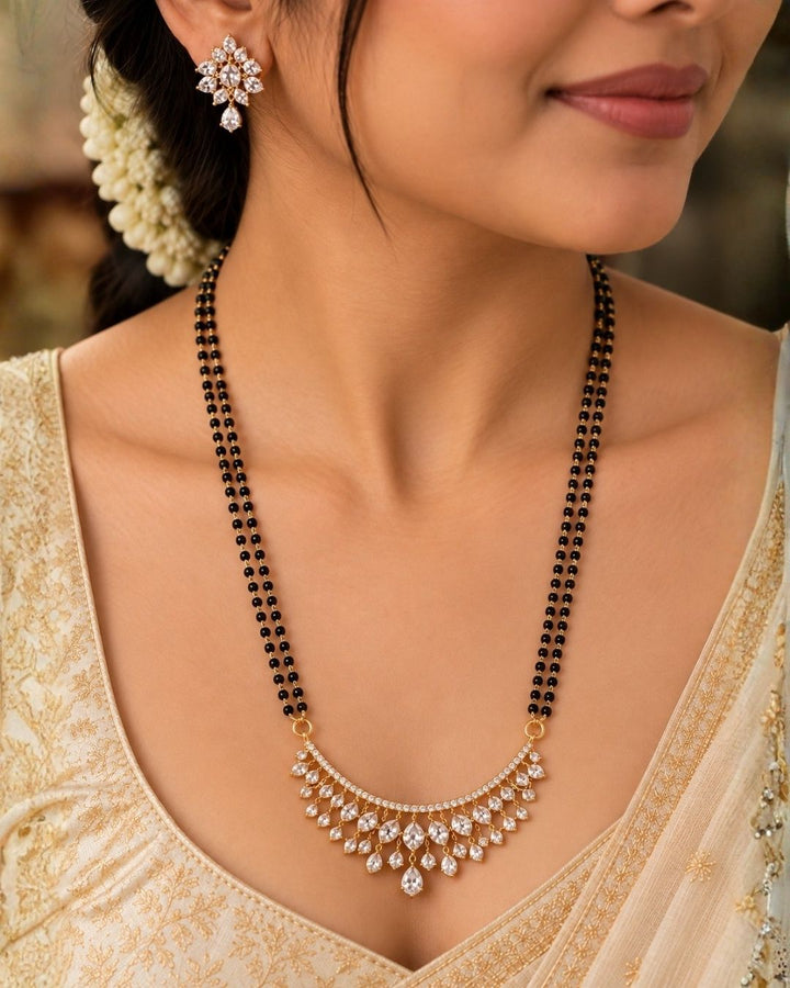 18K Gold Plated Zircon Diamond Mangalsutra set designed with a dual-strand black bead Nallapusalu chain and a half moon-shaped pendant. The pendant show rows of sparkling white zircon stones in a radiating fringe pattern, paired with matching cluster stone stud earrings for a luxurious, premium jewelry finish. Perfect for any ocaasions and dailywaer