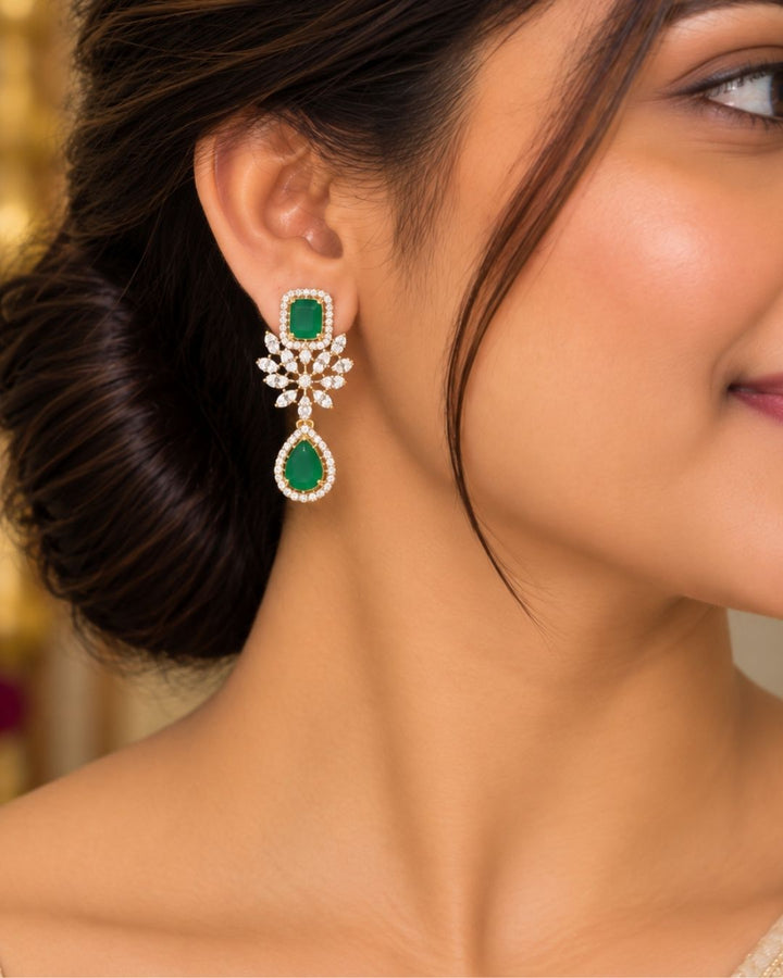 It centers an intricate, V-shaped floral bib pattern encrusted with glittering white American Diamonds (CZ stones), a prominent square emerald-green center stone, and a dangling teardrop emerald charm at the base. Includes a matching pair of elegant hanging earrings featuring a floral design with square and teardrop green stone accents. Perfect for parties and festive occasions with skin-friendly premium quality, comfortable fit,