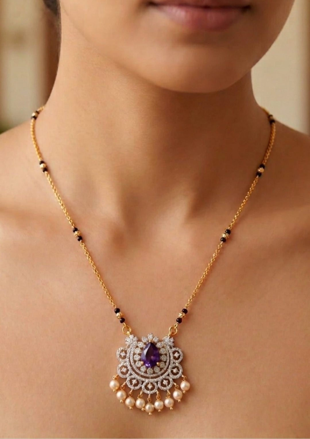 Gold necklace with a pendant featuring a purple gemstone on a Modern Zircon Diamond gold plated mangalsutra necklace The design features a delicate gold link chain transitioning into sections of traditional small black beads on both sides. The central pendant showcases a sleek, contemporary curved u-shape and a stylized butterfly or floral motif encrusted with sparkling white cubic zirconia stones. This elegant neckpiece perfect for daily wear, office wear, and casual outfits.person&