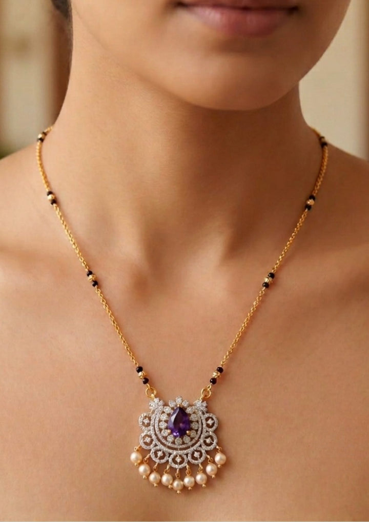 Gold necklace with a pendant featuring a purple gemstone on a Modern Zircon Diamond gold plated mangalsutra necklace The design features a delicate gold link chain transitioning into sections of traditional small black beads on both sides. The central pendant showcases a sleek, contemporary curved u-shape and a stylized butterfly or floral motif encrusted with sparkling white cubic zirconia stones. This elegant neckpiece perfect for daily wear, office wear, and casual outfits.person&
