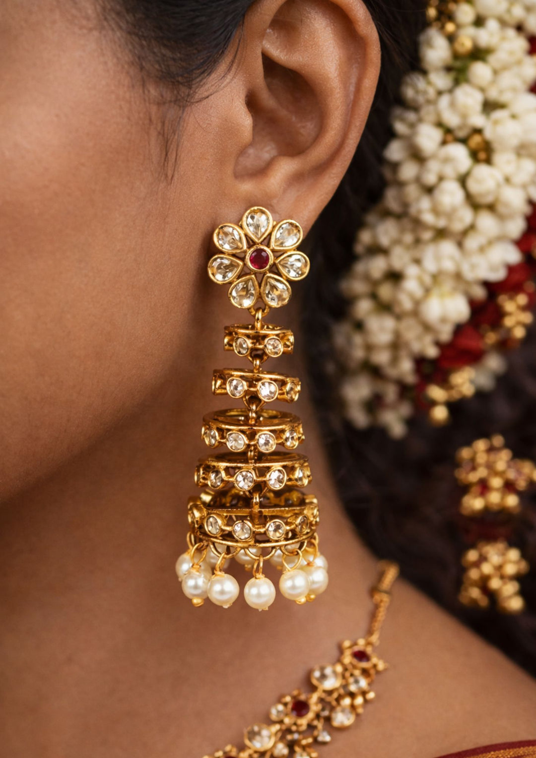 Close-up of a south indian woman wearing Gold Plated Traditional Jhumkas with dome-style buttalu pattern, colorful stone work, and pearl drops that give a grand bridal and festive jewelry look. The earrings come with lightweight comfort, sweat proof finish, anti-fade colour, smooth fitting, and are suitable for weddings, receptions, functions, and celebrations.