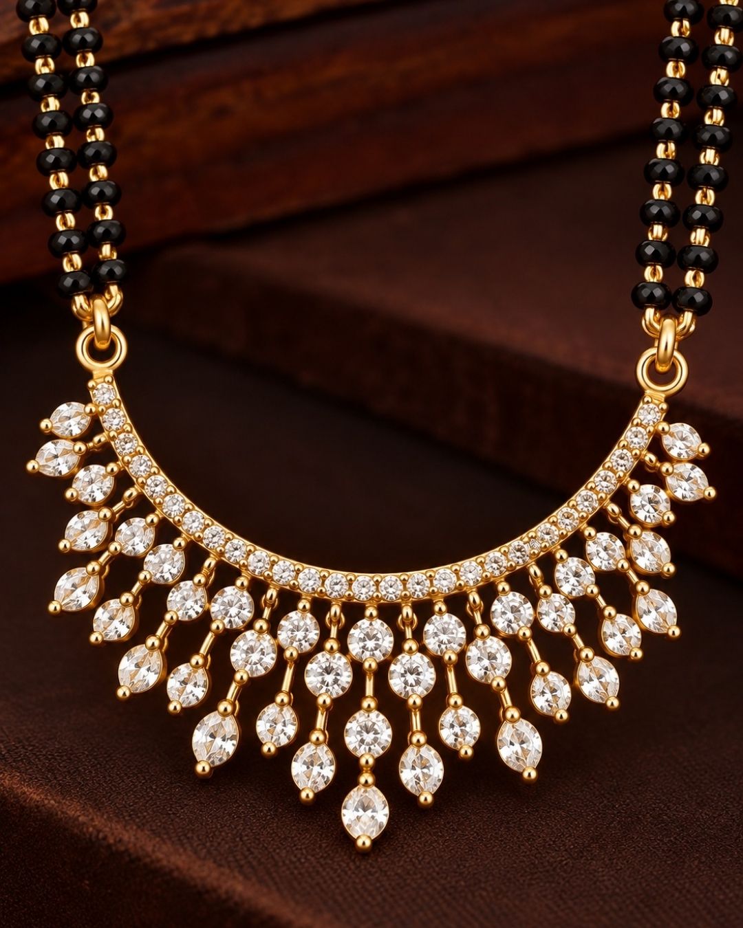 18K Gold Plated Zircon Diamond Mangalsutra set designed with a dual-strand black bead Nallapusalu chain and a half moon-shaped pendant. The pendant show rows of sparkling white zircon stones in a radiating fringe pattern, paired with matching cluster stone stud earrings for a luxurious, premium jewelry finish. Perfect for any ocaasions and dailywaer