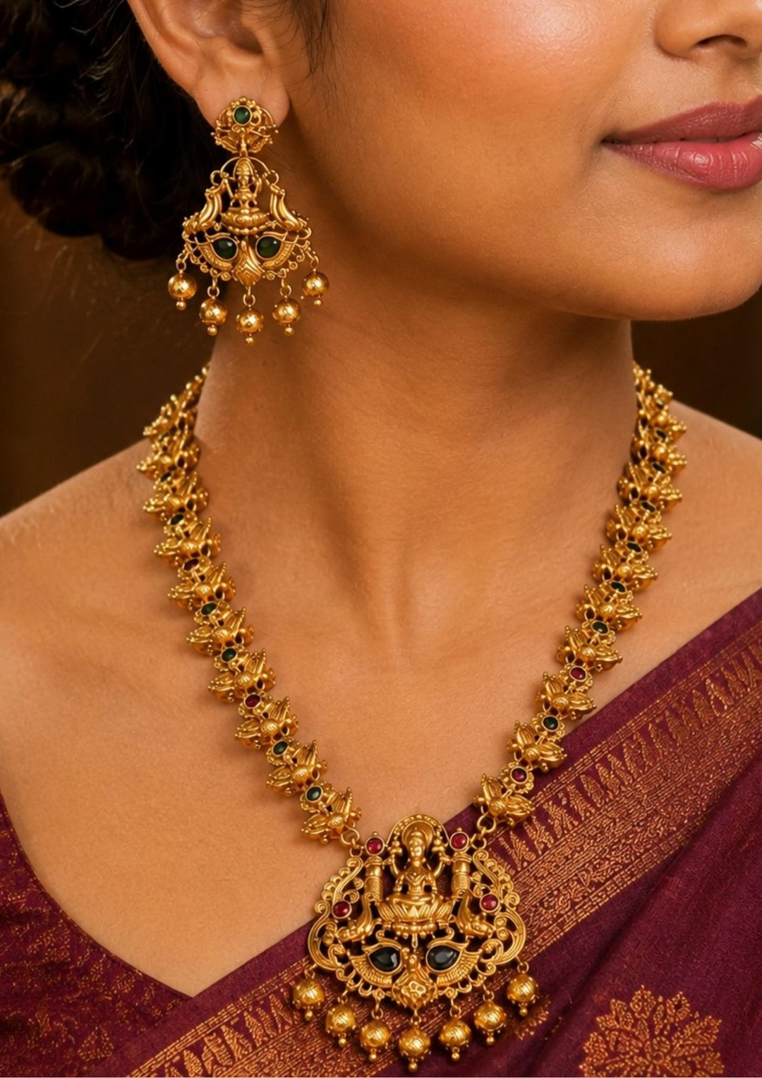 South Indian Women An exquisite antique gold-plated long temple haaram set featuring a prominent Goddess Laxmi pendant. The large, detailed pendant shows the deity seated on a lotus flanked by peacocks, encrusted with ruby-red and emerald-green stones, and finished with hanging textured gold bead balls. Perfect for for bridal and festive wear. It is anti tarnish and skin friendly