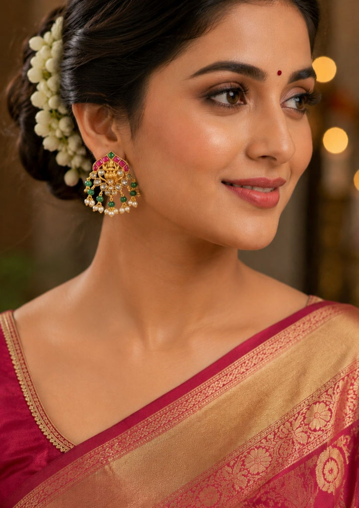 South Indian Women wearing A pair of antique temple-style gold-plated stud earrings intricately crafted with a central Goddess Laxmi motif seated on a lotus flower. The top edge is bordered with an arch of square-cut pink kemp stones and a single green accent, while the lower filigree tier is adorned with small white stones, hanging emerald-green round beads. Perfect for weddings and festive wear.