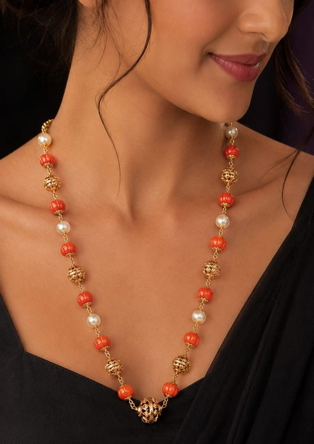 A close-up of a model wearing a long beaded necklace with orange coral-like beads, white pearl-like beads, and ornate gold-toned spacer beads, paired with a black top.